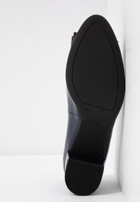 Chaussure habillée élégante à semelle noire, positionnée en angle contre un coin blanc, montrant les détails de la semelle et la marque "hōgl".