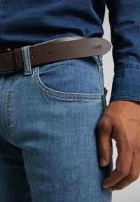 Jean en denim bleu avec une coupe ajustée, accompagné d'une ceinture en cuir brun. Gros plan sur la poche avant et la main reposant sur la cuisse.
