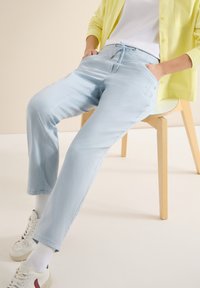 Personne assise sur une chaise en bois clair, portant un pantalon bleu clair à cordon, des baskets blanches, des chaussettes blanches, une chemise blanche et un cardigan jaune.