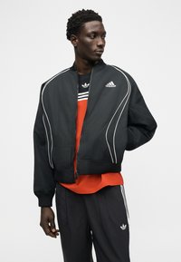 Νέος άνδρας με μαύρο μπουφάν Adidas και παντελόνι με λευκές ρίγες, φοράει πορτοκαλί μπλούζα από κάτω, στέκεται απέναντι σε απλό φόντο.