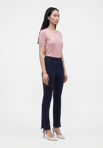Femme debout portant un haut rose ajusté à manches courtes, un pantalon taille haute bleu marine foncé avec des fentes latérales et des escarpins beiges à bout pointu.
