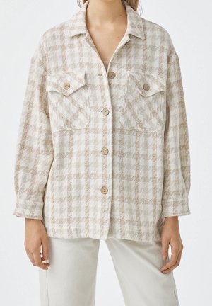 Veste-chemise à carreaux beige et blanche avec deux poches poitrine et des boutons marron, portée avec un pantalon de couleur claire.