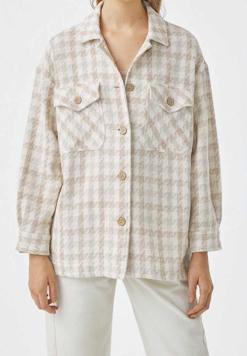 Veste-chemise à carreaux beige et blanche avec deux poches poitrine et des boutons marron, portée avec un pantalon de couleur claire.