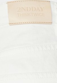 Witte stof met textuur, voorzien van een beige leren label met daarop subtiel gedrukt "2NDDAY" en "THINKTWICE".