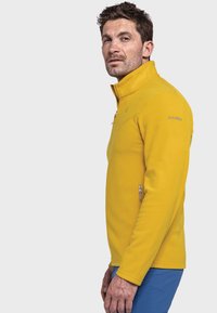 Gelbe Fleecejacke mit hohem Kragen, Reißverschlusstaschen und Logodetail auf der Schulter; kombiniert mit blauen Hosen.