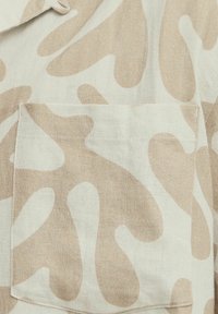 Un tissu à motif camouflage beige présente une surface texturée avec une poche à rabat. Le matériau semble léger et respirant.