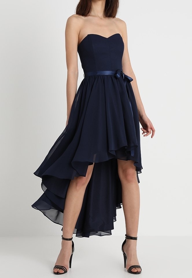 Robe bustier bleu marine à ourlet asymétrique avec jupe à couches et nœud en satin à la taille, portée avec des sandales noires à talons hauts et bride à la cheville.