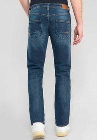 Vaqueros de mezclilla azul oscuro con un ajuste slim, que cuentan con un parche de cuero marrón en la parte trasera, dos bolsillos traseros de parche y sutiles detalles de costura.