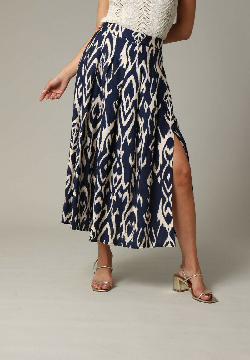 Jupe marine avec un motif ikat beige, design plissé et fente sur le côté. Portée avec des sandales à talons blocs dorés.