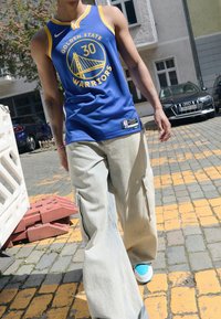 Maglia da basket blu con accenti gialli e logo. Pantaloni cargo larghi di colore chiaro con tasche laterali. Sneakers blu con suole bianche.