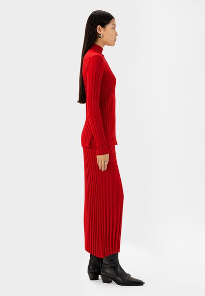 Rotes geripptes Strickkleid mit hohem Kragen und seitlichen Schlitzen, kombiniert mit schwarzen Knöchelstiefeln. Verfügt über vertikale Struktur und figurbetonte Silhouette.