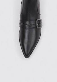 Chaussure en cuir noir à bout pointu avec une bride décorative ornée d'œillets métalliques et d'une boucle en argent sur le devant.