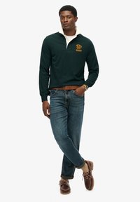 Camiseta de rugby de manga larga color verde oscuro con cuello blanco, que presenta el texto "RUGBY" en color naranja. Se lleva con jeans azules y zapatos marrones de cordones.