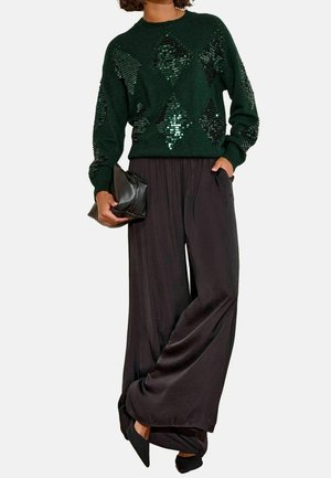 Osoba ubrana w ciemnozielony sweter z cekinami w diamentowy wzór, szerokie czarne spodnie, czarne spiczaste buty, trzymająca czarną kopertówkę.