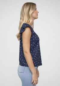 Tmavě modrý top s bílým vzorem srdcí, krátkými volánkovými rukávy a volným střihem, doplněný světle modrými přiléhavými džínami.