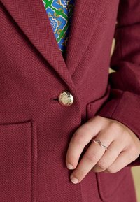 Burgundi blazer, milleksest mustrist, millel on läikiv kuldne nupp. Krae on sälguline ja värviline mustriga kangas on nähtav allpool.