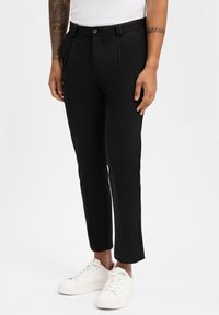 Schwarze maßgeschneiderte Hose mit Falten, aus glattem Stoff. Getragen mit weißen Sneakers, mit Knopfverschluss und Gürtelschlaufen.