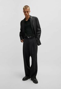 Schwarze Lederjacke über einem dunklen Polo-Shirt, kombiniert mit lockeren schwarzen Hosen und schwarzen Schuhen. Schlichte, klare Linien und minimale Details.
