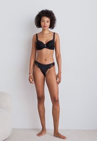 Schwarzer Spitzen-BH mit Unterkonstruktion und einem kleinen silbernen Akzent; dazu passende schwarze Spitzenunterhose mit weicher Textur und minimalen Designdetails.