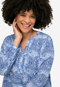 Blusa azul con estampado, escote en V, mangas largas y detalle de botones. Presenta intrincados diseños geométricos en varios tonos de azul.