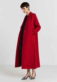 Langer roter Mantel mit maßgeschneiderter Silhouette, ein Knopfverschluss und zwei seitlichen Taschen, kombiniert mit einem schwarzen Kleid und Zebramuster-Absätzen.
