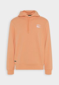 Sweatshirt à capuche en tissu doux pêche avec poche kangourou, logo Lacoste blanc et logo d'accent vert à l'ourlet. Dispose d'un col avec cordon de serrage.