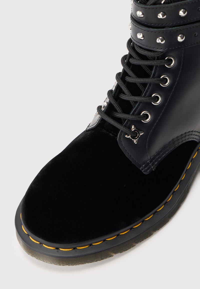 Black ankle boot featuring a glossy upper, velvet toe, decorative studs, and yellow stitching along the sole. Laced design with metal eyelets.