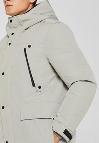 Giacca isolante grigio chiaro con cappuccio, tasca laterale con zip e dettagli in hardware neri. Presenta una texture liscia e un design moderno.