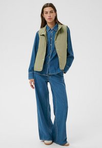 Denim-Outfit besteht aus einer hellgrünen Weste über einem blauen Hemd mit Knöpfen und weitem Jeanshose, die eine harmonische Farbpalette und eine lässige Passform zeigen.