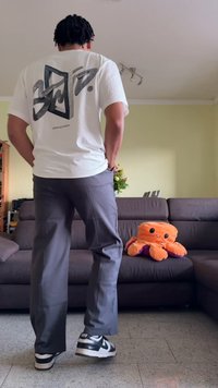 T-shirt branca com estampado gráfico nas costas, calças cinzentas e sapatilhas pretas. Um boneco de polvo laranja em peluche repousa num sofá escuro.