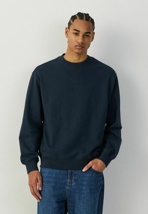 Junger Mann mit geflochtenem Haar, der einen marineblauen Sweatshirt mit Rundhalsausschnitt und blaue Jeans trägt, steht vor einem einfachen hellen Hintergrund.
