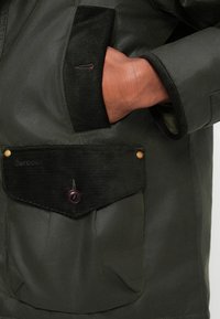 Jachetă verde închis cu un buzunar din catifea cu un nasture mov. Exterior neted cu un luciu subtil, evidențiind materialul său durabil.