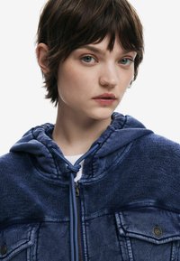 Dunkelblaue Jeansjacke mit strukturierter Kapuze, zwei Brusttaschen und Reißverschluss. Besitzt Kordelzüge und kontrastierende Stoffdetails.