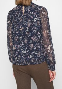 Blusa floreale color navy con maniche lunghe in tessuto trasparente, apertura a chiave con bottone sul retro e polsini elasticizzati. Presenta motivi floreali rosa e verdi su uno sfondo scuro.