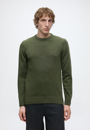 Jeune homme aux cheveux blonds bouclés portant un pull en tricot vert uni et un pantalon foncé, debout devant un fond blanc.