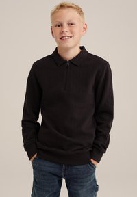 Schwarzer Pullover mit Reißverschlusskragen, strukturiertem Stoff, gerippte Bündchen und Saum. Über blauen Jeans getragen, mit einem lockeren Design und Schnitt.