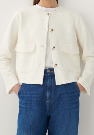 Frau trägt eine weiße, strukturierte, geknöpfte Jacke mit Vordertaschen über einem weißen Top, kombiniert mit hoch taillierten blauen Jeans, Hände in den Taschen.