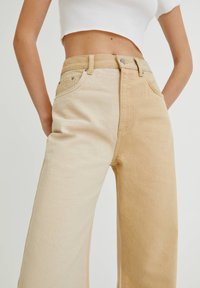 Gros plan sur une personne portant un jean taille haute à jambes larges bicolore beige et tan, associé à un haut court en coton côtelé blanc, mains derrière le dos.