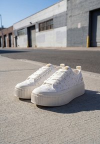 Λευκά sneakers με πλατφόρμα και μικρά μοτίβα μαργαρίτας σε τσιμεντένιο πεζοδρόμιο κοντά σε βιομηχανικά κτίρια κάτω από καθαρό ουρανό.