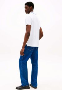 Witte poloshirt met een navy kraag, gecombineerd met blauwe jeans met rechte pijpen en zwarte schoenen. Zichtbare naden en zijsplits in het shirt.