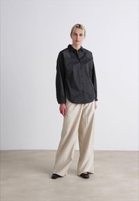Cămașă cu dungi negre și guler clasic, asortată cu pantaloni largi bej și pantofi negri. Fundal neutru, stil minimalistic.