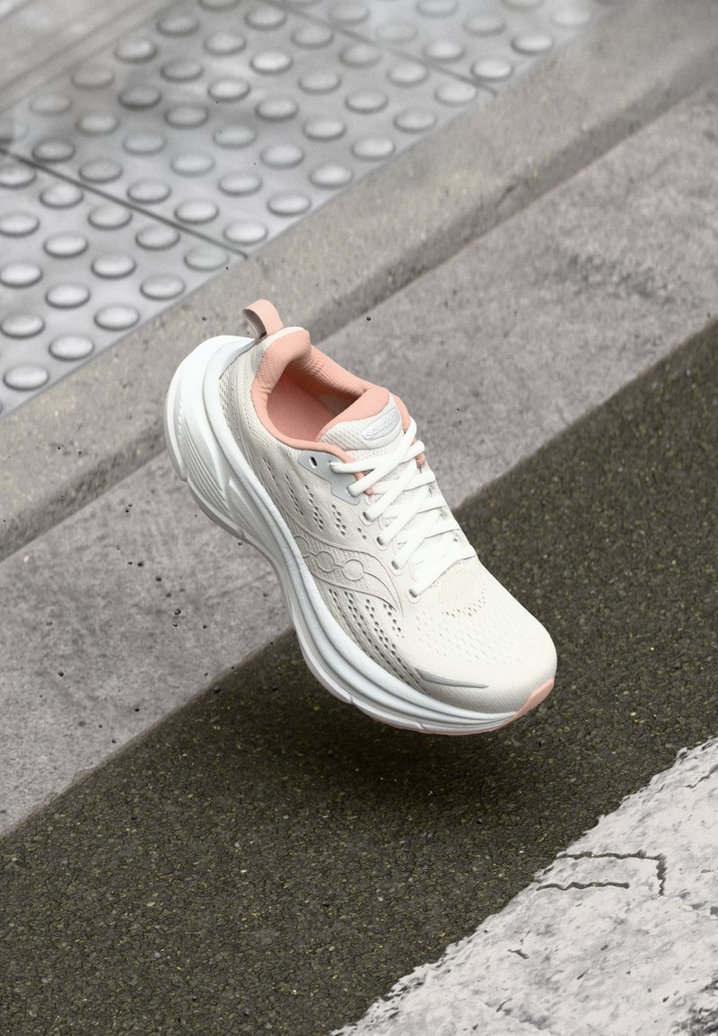 Białe sportowe buty z teksturowaną powierzchnią, różowymi akcentami, sznurowanym designem i grubą amortyzacją. Wyraźne perforacje i szczegóły z logo.