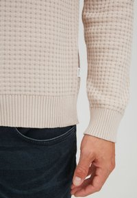 Pull en maille beige clair avec un motif texturé, poignets et ourlet côtelés. Comprend une petite étiquette de marque sur le côté. Associé à un jean foncé.