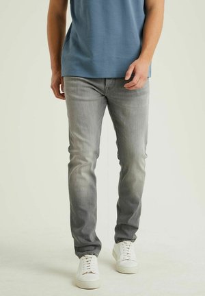 Man wearing faded gray jeans, white sneakers, and a blue shirt, standing against a plain light background.