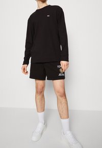 Svart långärmad tröja med liten logga, tillsammans med svarta shorts med en logga och text. Bärs med vita strumpor och sneakers.