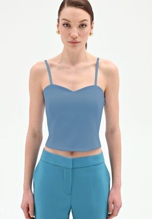 Femme portant un haut court bleu sans manches et un pantalon taille haute assorti, avec des boucles d'oreilles dorées en forme d'anneau, face à l'avant sur un fond blanc.