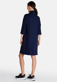 Vestido azul marino hasta la rodilla con cuello alto y mangas tres cuartos. Lleva zapatillas negras con suela blanca y logo.