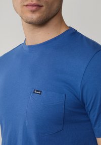 Niebieska koszulka z krótkim rękawem z gładkiej mieszanki bawełny, z okrągłym dekoltem i naszywaną kieszenią z metką po lewej stronie na piersi.