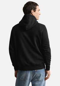 Sort hættetrøje med hætte, der har en glat tekstur. Båret over lyseblå jeans, der viser et enkelt, klassisk design.