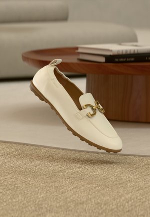 Kremkrāsas ādas loaferi ar zelta sprādzes detaļu un gumijas zolīti, novietoti uz strukturēta bēša paklāja ar koka kafijas galdiņu un grāmatām fonā.